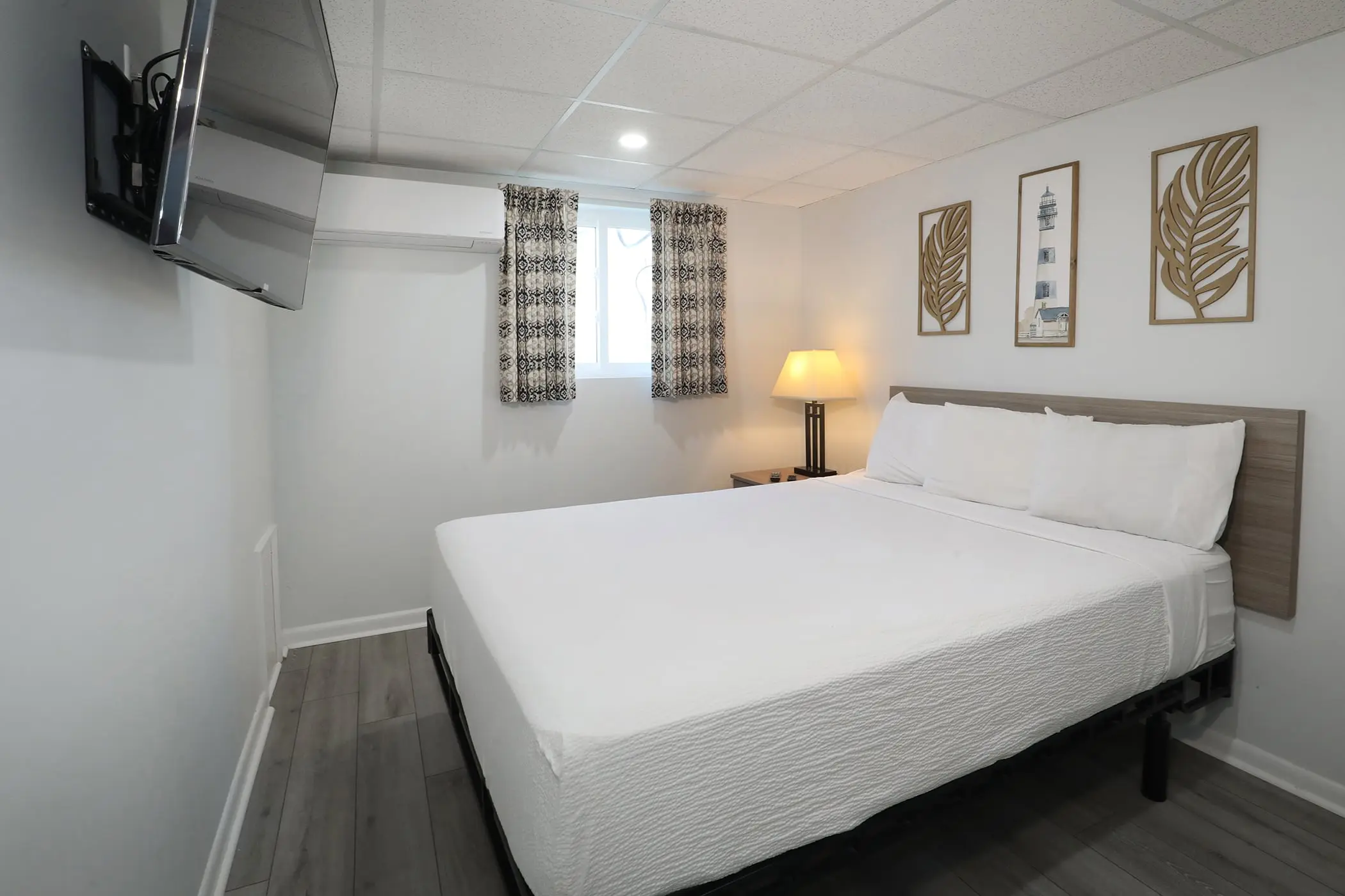 One bedroom of a Type 8 Two-Bedroom Unit at the Matador: a single large bed, bedside lamp, wall-mounted TV and shore-themed wall art