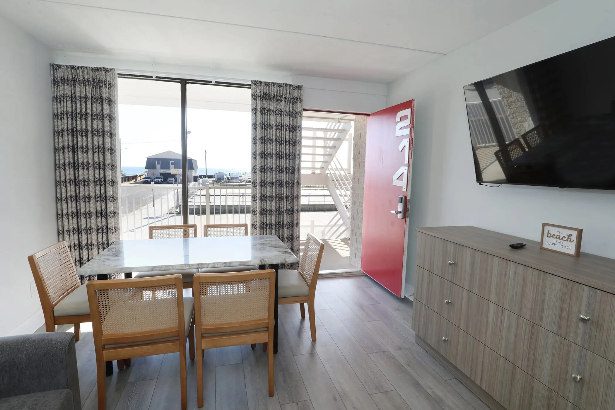 The dining table and rattan-back chairs in a Type 8 Two-Bedroom Unit at the Matador, beside the sliding glass door and the red front door of unit 214